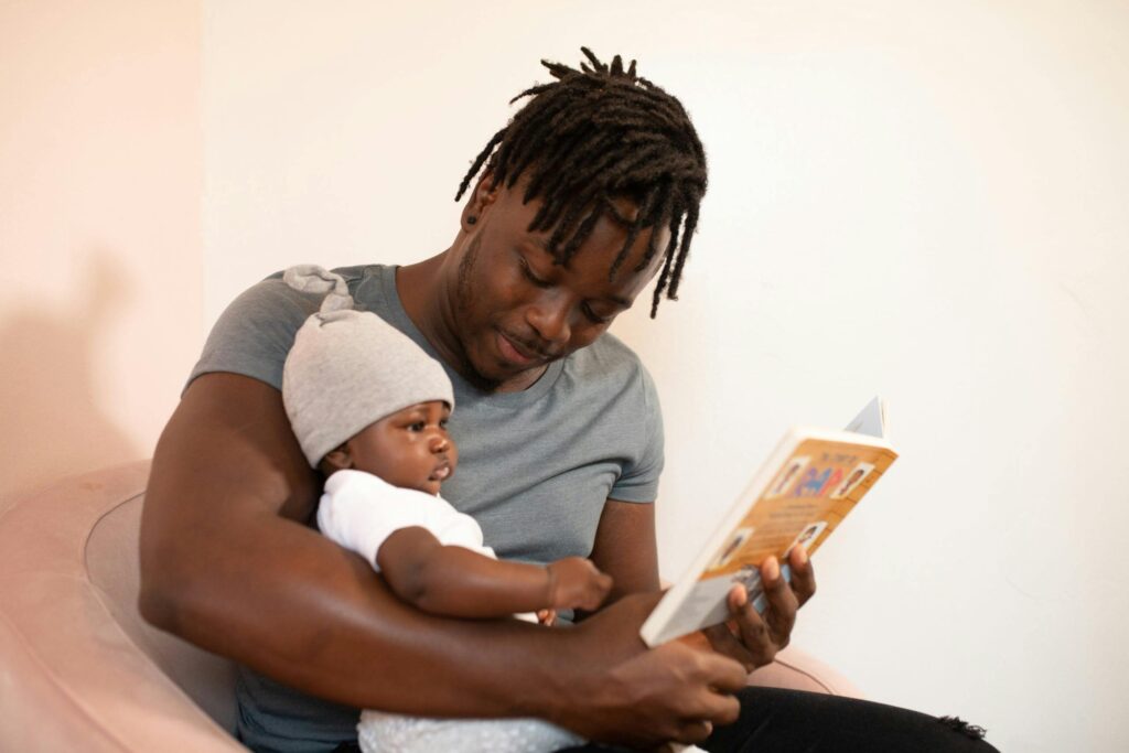 pexels-photo-3536630-3536630 A father with dreadlocks reads a book to his baby, creating a bonding moment.